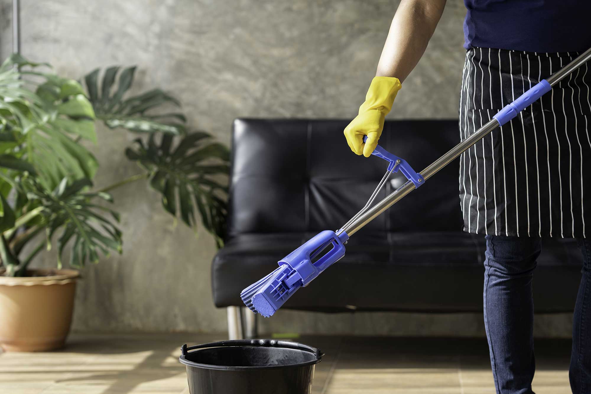A person in a striped apron and yellow gloves prepares to clean with a blue mop over a black bucket, surrounded by greenery.