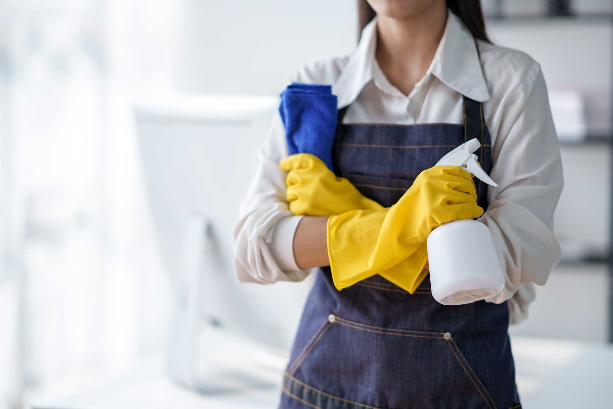A person wearing yellow rubber gloves holds a spray bottle and a blue cloth, ready for cleaning tasks in a bright, modern space.