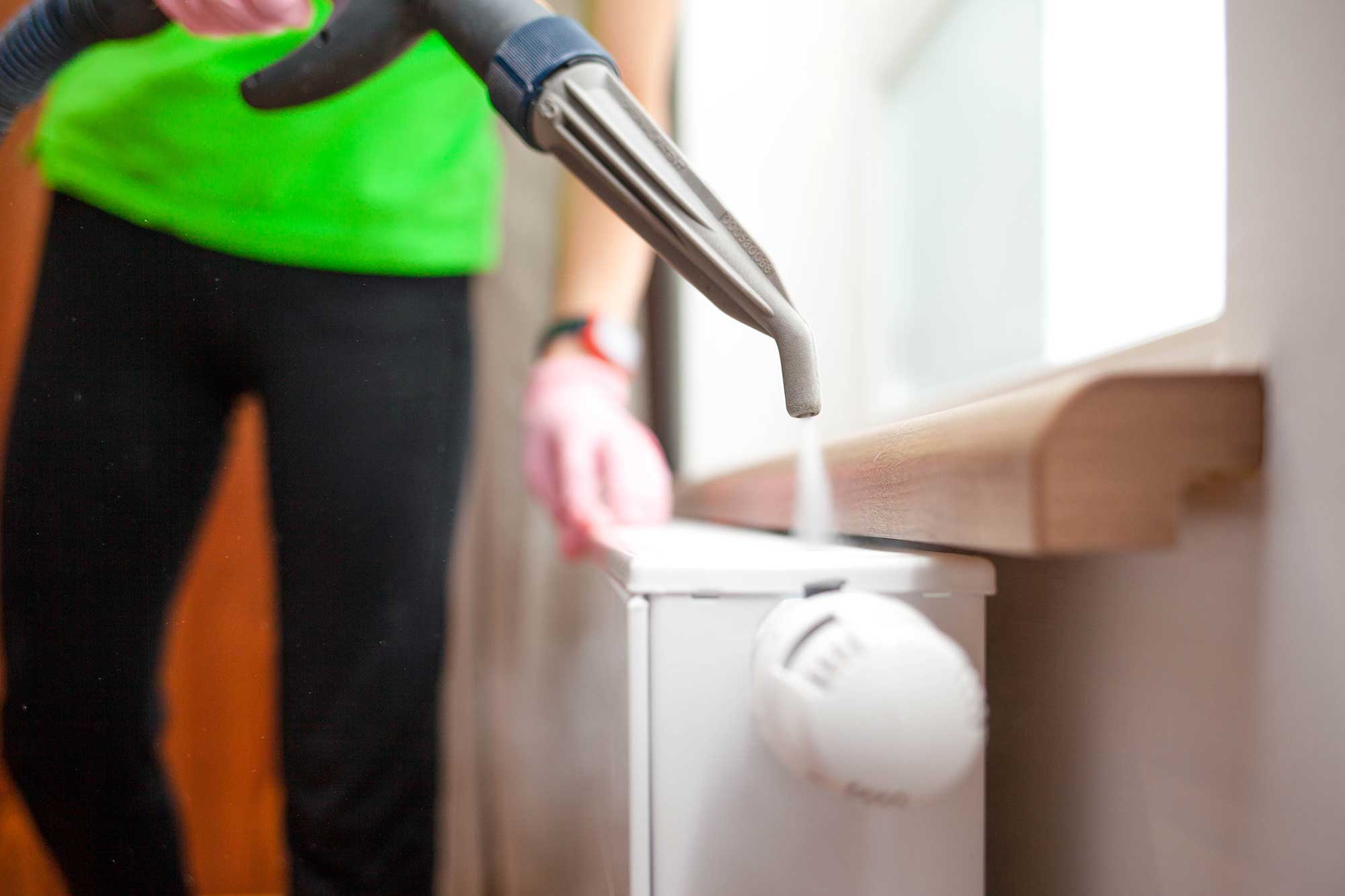 A person in a green shirt cleans a white radiator with a steam cleaner, with steam visibly rising from the nozzle.
