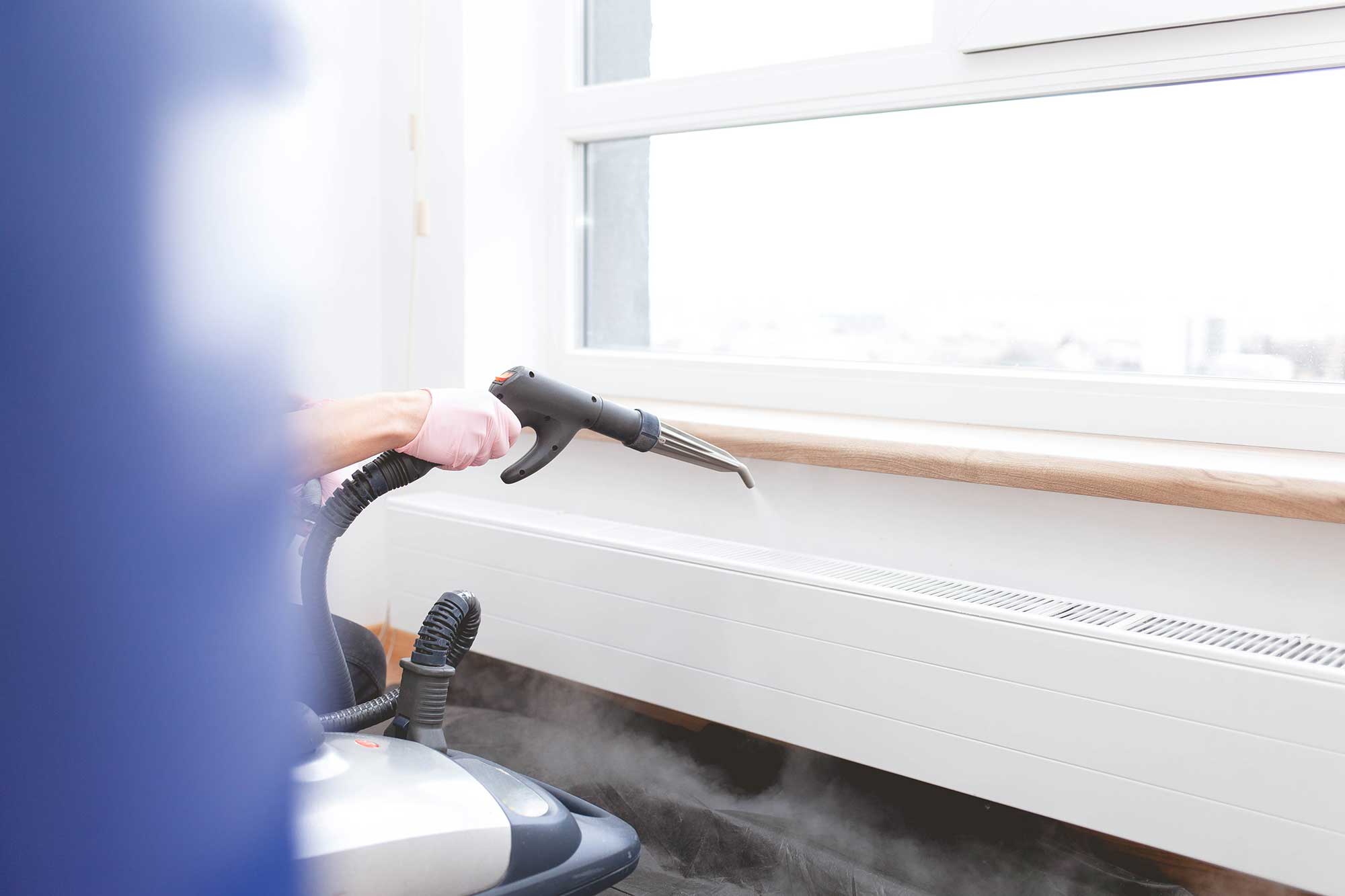 A person wearing pink gloves uses a steam cleaner to sanitize a windowsill, with steam rising and sunlight flooding the room.