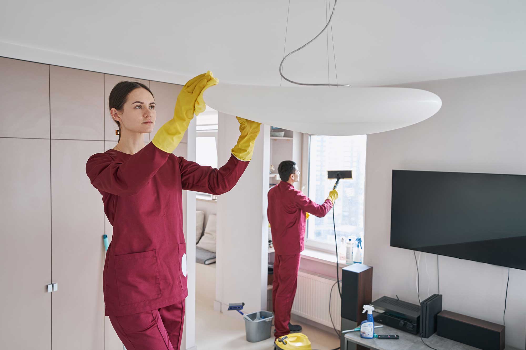 A person in maroon cleaning attire and yellow gloves is adjusting a ceiling light, while another person cleans a window nearby.
